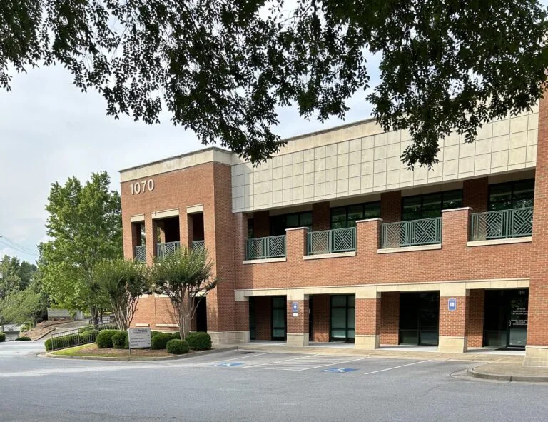 A two-story brick building displays the number “1070” on the upper left facade, with large windows and a balcony featuring green railings. In front, a landscaped area with trees, bushes, and marked parking spaces—including accessible spots—creates a clean and welcoming exterior under an overcast sky.